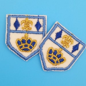 Könnte beinhalten: Zwei weiße Stoffpatches mit blauem Besatz und goldener Stickerei. Die Patches zeigen ein Kronendesign und einen stilisierten Buchstaben C.