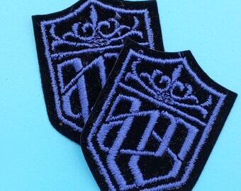 May include: Two black felt patches with purple embroidered designs. The designs are intricate and feature a crown and other decorative elements.