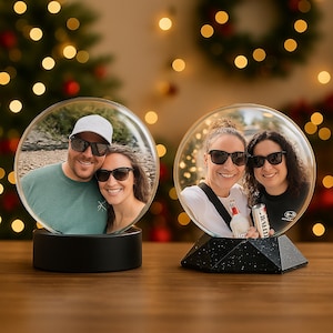 Può includere: Due globi fotografici illuminati con foto di persone all'interno. Uno ha una base nera rotonda, l'altro una base geometrica nera. Lo sfondo presenta luci natalizie e una ghirlanda, suggerendo un tema festivo.
