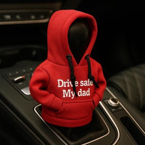 May include: A red hoodie car accessory with the text "Drive safe My dad" in white. The hoodie has a black drawstring and is placed on a car's gear shift. The accessory is designed to fit over a car's gear shift.
