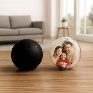 May include: A black, spherical speaker with a smiling face design is positioned beside a clear glass sphere. Inside the sphere, a family of three is pictured in front of a Christmas tree. The objects are on a wooden surface.