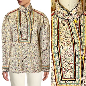 May include: A white cotton shirt with a colorful geometric pattern. The shirt has a high collar and long sleeves. The pattern is made up of red, green, yellow, and black stripes.