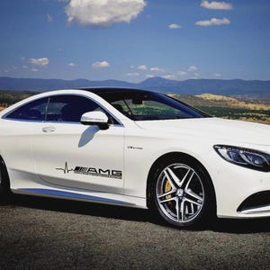 May include: White coupe car with black detailing, parked on a road. The car has a black roof and silver alloy wheels. A black AMG logo with "DRIVING PERFORMANCE" is on the side. The background features mountains and a blue sky with clouds.