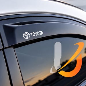 Può includere: Un deflettore antipioggia per finestrino auto in plastica nera con il logo Toyota stampato in bianco sulla superficie. Il deflettore è montato lungo il bordo superiore del telaio del finestrino per deviare pioggia e vento durante la guida.