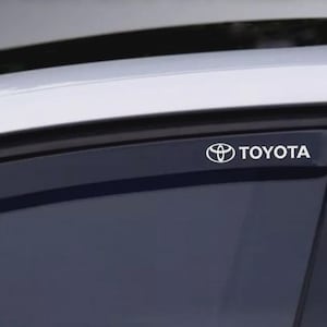 May include: A close-up of a car window with a white Toyota logo and text. The logo is a stylized emblem with the brand name in a sans-serif font. The window is tinted, and the car's roof is visible.