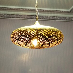 Op de afbeelding: Een gouden metalen hanglamp met een ronde, opengewerkte kap. De kap heeft een geometrisch patroon van kleine cirkels en grotere, bloemvormige vormen. De lamp hangt aan een ketting.