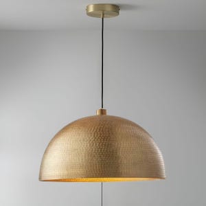 May include: A gold-coloured, dome-shaped pendant light fitting. The light has a textured surface and hangs from a black cord. The fitting is attached to a gold-coloured ceiling mount.