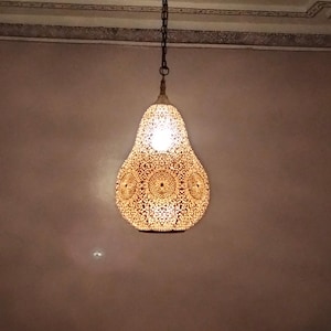 Op de afbeelding: Een peervormige, sierlijke hanglamp met een warme, gloeiende binnenkant. De buitenkant heeft een gedetailleerd, geperforeerd patroon en hangt aan een ketting. Het licht hangt tegen een neutraal getinte muur.