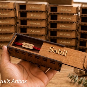 May include: A rectangular wooden sliding box featuring the name Sahil engraved on the lid and a red string bracelet with a small wooden tag also inscribed with the name Sahil inside. The box has a dark brown finish with visible wood grain.