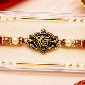 Handgefertigtes Ik Onkar Rakhi: Antikgoldfarben, Perle, rubinrote Kristallakzente