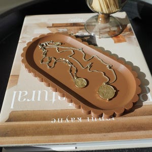 May include: A brown oval-shaped tray with scalloped edges, featuring two gold necklaces with circular pendants. The tray is resting on a book with the text "omni kayne" visible.