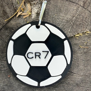 Puede incluir: Adorno de balón de fútbol en blanco y negro con el texto "CR7" en el centro. El adorno es redondo con un lazo de metal plateado para colgar. El diseño del balón de fútbol presenta pentágonos blancos y hexágonos negros.