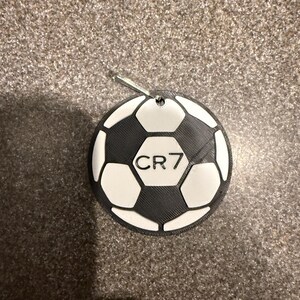 May include: A black and white soccer ball ornament with the text "CR7" in the center. The ornament has a silver loop for hanging. The soccer ball design is made up of black pentagons and white hexagons.