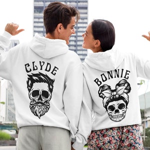 Könnte beinhalten: Zwei weiße Hoodies mit schwarzen Totenkopf-Grafiken und dem Text "Clyde" und "Bonnie" auf der Rückseite.