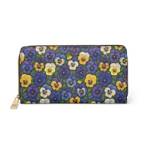 Op de afbeelding: Een portemonnee met ritssluiting met een bloemenpatroon van blauwe, gele en witte viooltjes op een donkergroene achtergrond. De portemonnee heeft een goudkleurige ritssluiting en trekker.