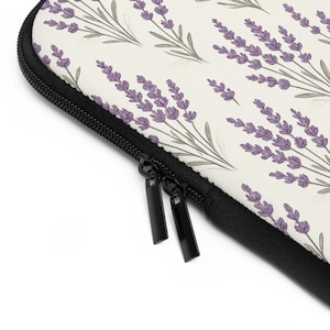 Puede incluir: Una funda para portátil con un fondo crema y un patrón repetido de ramitas de lavanda en morado y gris. Tiene una cremallera negra y un borde negro.