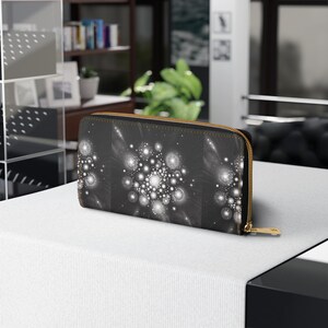 Pode incluir: Carteira preta com zíper dourado e design abstrato branco e cinza. A carteira é retangular e parece ser feita de um material texturizado. O design apresenta aglomerados de círculos e pontos brancos.