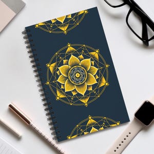 May include: A spiral-bound notebook with a navy blue cover featuring a gold geometric floral design. The design includes a central flower with a detailed eye-like center and geometric patterns. The notebook is on a white surface.