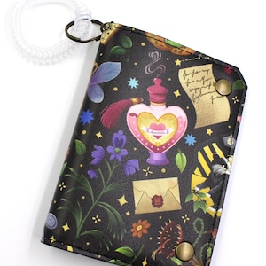 May include: Black wallet with a whimsical design featuring a heart-shaped potion bottle, flowers, and stars. The wallet has a wrist coil and a metal clasp. The design includes an envelope with a wax seal and a scroll.