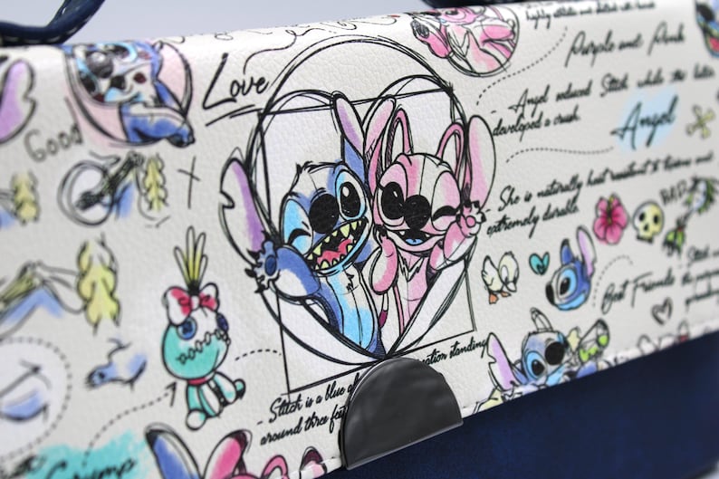 May include: A white handbag with a navy blue base and handle. The flap features a cartoon illustration of Stitch and Angel, with handwritten text and sketches. The design includes the words "Love" and "Angel".