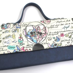 May include: A navy blue and cream-colored clutch featuring a cartoon illustration of Stitch and Angel. The clutch has a dark blue handle and a magnetic closure. The design includes hearts, flowers, and text.