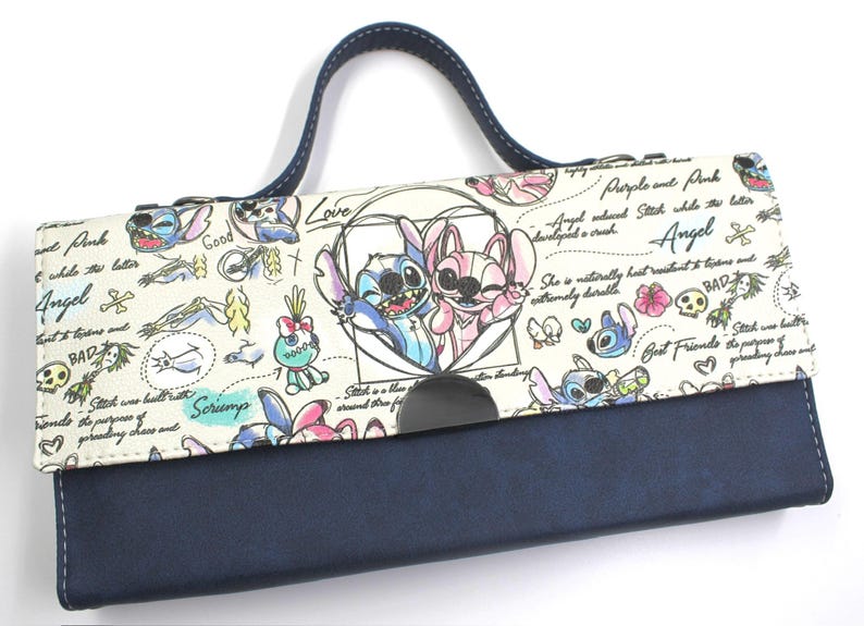 May include: A navy blue and cream-colored clutch featuring a cartoon illustration of Stitch and Angel. The clutch has a dark blue handle and a magnetic closure. The design includes hearts, flowers, and text.