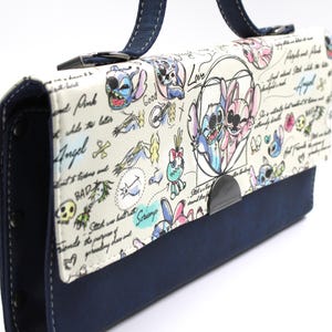 May include: A white handbag with a navy blue base and handle. The bag features a cartoon design with the characters Stitch and Angel, along with handwritten text and doodles. The bag has a metal clasp closure.