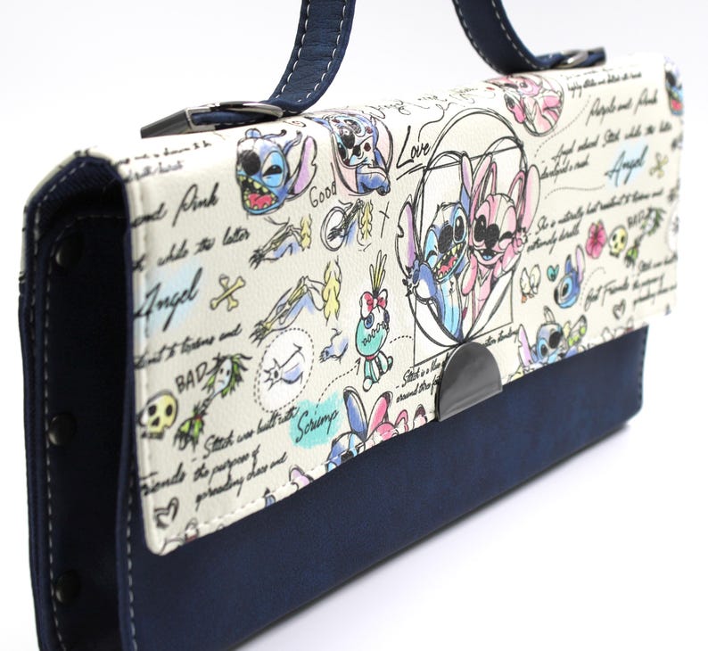 May include: A white handbag with a navy blue base and handle. The bag features a cartoon design with the characters Stitch and Angel, along with handwritten text and doodles. The bag has a metal clasp closure.