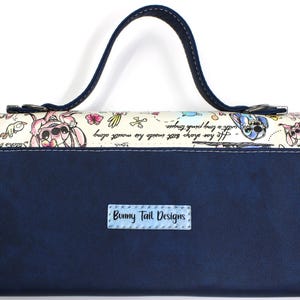 May include: A navy blue clutch with a handle, featuring a whimsical design. The top section has a cream background with cartoon characters and text. The bottom is solid navy blue with a small label that reads "Bunny Tail Designs".