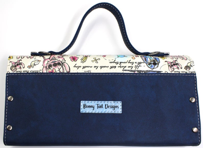 May include: A navy blue clutch with a handle, featuring a whimsical design. The top section has a cream background with cartoon characters and text. The bottom is solid navy blue with a small label that reads "Bunny Tail Designs".