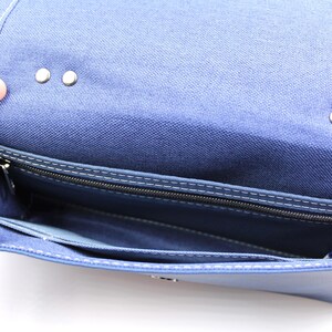 May include: Open view of a blue fabric purse with a flap closure. The interior reveals a zippered pocket and several compartments. Silver-colored rivets secure the flap. White stitching accents the edges.