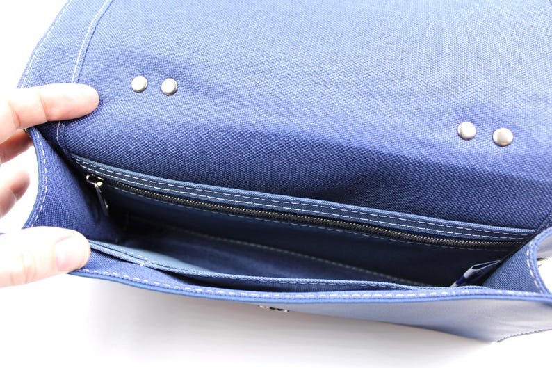 May include: Open view of a blue fabric purse with a flap closure. The interior reveals a zippered pocket and several compartments. Silver-colored rivets secure the flap. White stitching accents the edges.