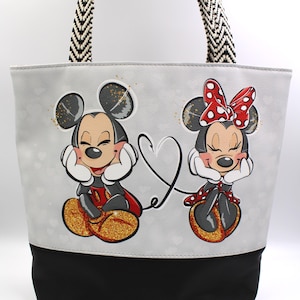 May include: Gray tote bag featuring Mickey and Minnie Mouse. Mickey is wearing red shorts and a black vest, while Minnie wears a red polka dot dress. The bag has a black base, a white and black patterned strap, and a heart design.