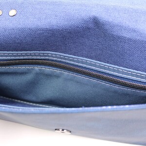 May include: Close-up of a blue canvas bag with a zippered pocket. The bag has a flap closure with metal rivets and a silver clasp. White stitching accents the seams and pocket.