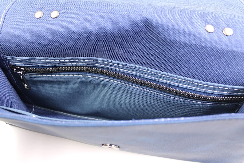May include: Close-up of a blue canvas bag with a zippered pocket. The bag has a flap closure with metal rivets and a silver clasp. White stitching accents the seams and pocket.