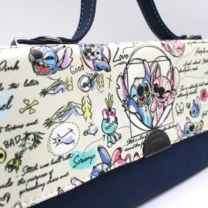 May include: A white and navy blue handbag featuring cartoon illustrations of Stitch and Angel. The bag has a navy blue handle and a flap with drawings and text, including the words "Angel" and "Love".