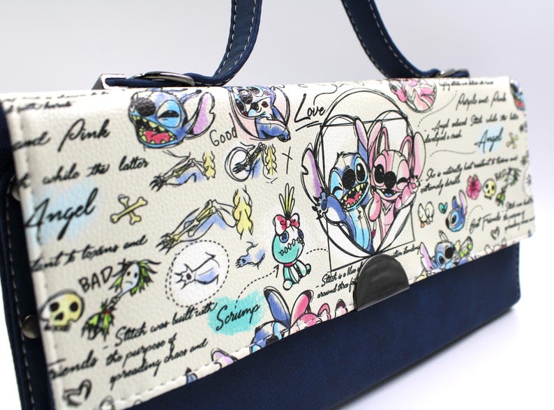 May include: A white and navy blue handbag featuring cartoon illustrations of Stitch and Angel. The bag has a navy blue handle and a flap with drawings and text, including the words "Angel" and "Love".