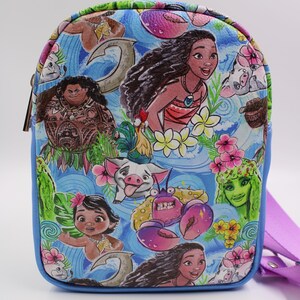 Puede incluir: Un bolso bandolera azul y morado con un estampado colorido con personajes de la pel&iacute;cula de Disney Moana. El estampado incluye a Moana, Maui, Pua el cerdo, Hei Hei el gallo y Tamatoa el cangrejo.