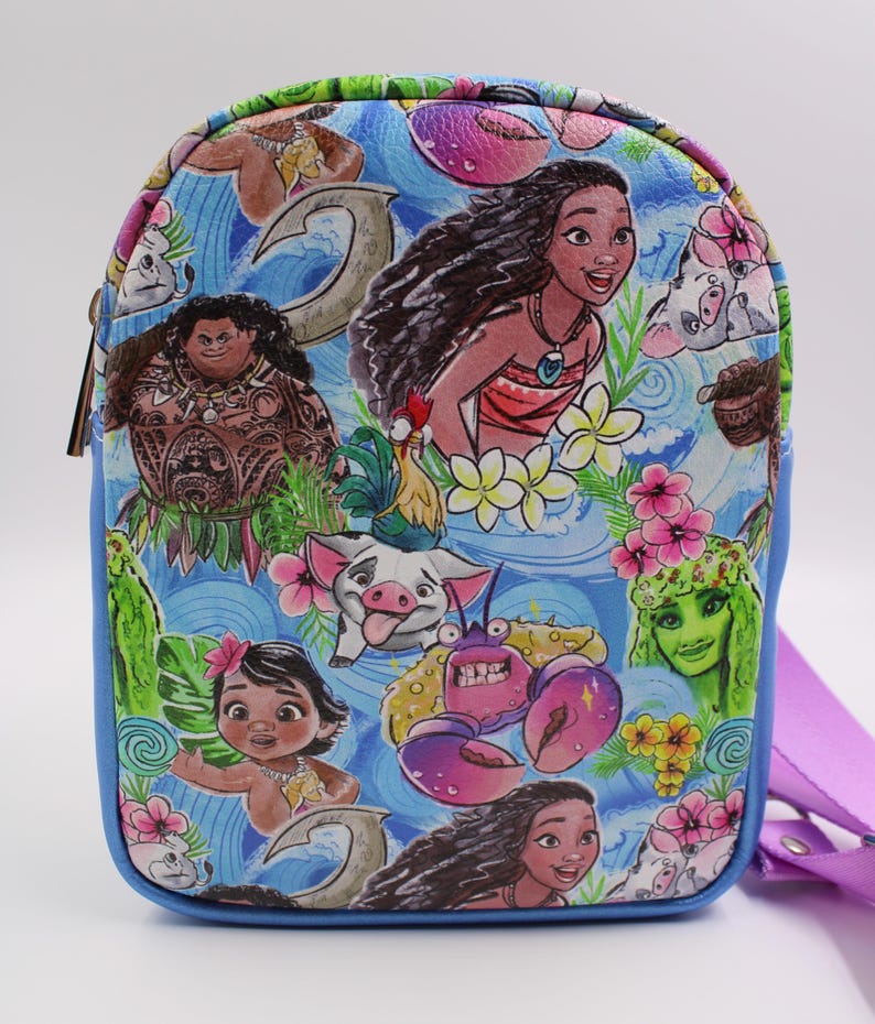 Puede incluir: Un bolso bandolera azul y morado con un estampado colorido con personajes de la pel&iacute;cula de Disney Moana. El estampado incluye a Moana, Maui, Pua el cerdo, Hei Hei el gallo y Tamatoa el cangrejo.