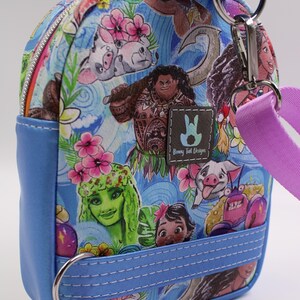 Puede incluir: Una peque&ntilde;a mochila azul con un estampado colorido con personajes de la pel&iacute;cula animada Moana. El estampado incluye a Moana, Maui, Pua el cerdo, Hei Hei el gallo y Te Fiti. La mochila tiene cierre de cremallera, un asa superior y una correa desmontable.