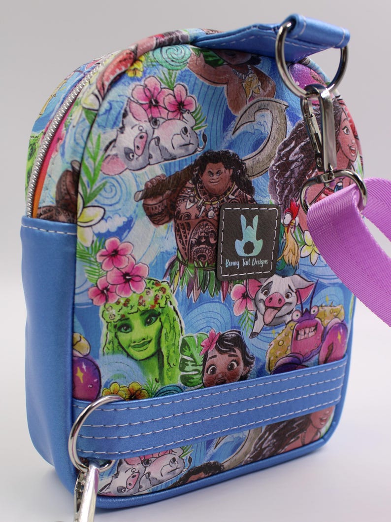 Puede incluir: Una peque&ntilde;a mochila azul con un estampado colorido con personajes de la pel&iacute;cula animada Moana. El estampado incluye a Moana, Maui, Pua el cerdo, Hei Hei el gallo y Te Fiti. La mochila tiene cierre de cremallera, un asa superior y una correa desmontable.