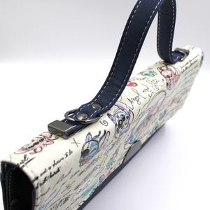May include: A white clutch with a navy blue handle and accents. The clutch features a colorful cartoon design with the words "Angel and Stitch" and other handwritten text. The handle is attached with a silver metal clasp.