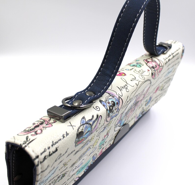May include: A white clutch with a navy blue handle and accents. The clutch features a colorful cartoon design with the words "Angel and Stitch" and other handwritten text. The handle is attached with a silver metal clasp.