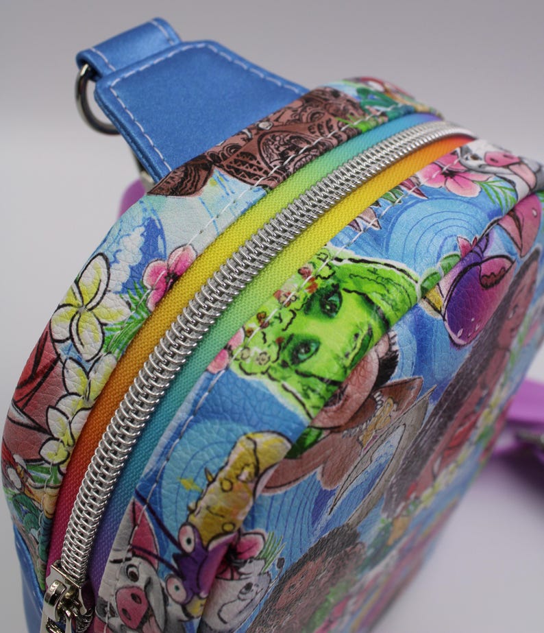 Puede incluir: Una peque&ntilde;a mochila azul con una cremallera arco&iacute;ris y un estampado colorido de personajes de dibujos animados y flores.