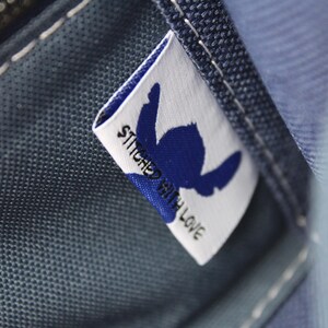 May include: Close-up of a blue fabric with a white label. The label features a blue silhouette and the words "STITCHED WITH LOVE". The fabric has visible stitching and a zipper.