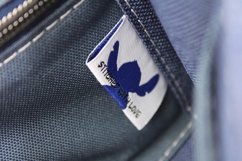 May include: Close-up of a blue fabric with a white label. The label features a blue silhouette and the words "STITCHED WITH LOVE". The fabric has visible stitching and a zipper.