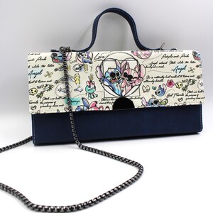 May include: A navy blue and cream-colored handbag with a chain strap and a top handle. The flap features a cartoon illustration of two characters in a heart, with the words "Love", "Angel", and "BAD" in the design. The bag is approximately 10 inches wide.