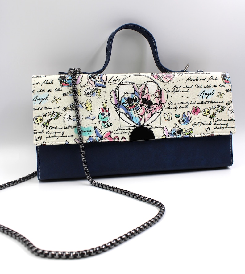 May include: A navy blue and cream-colored handbag with a chain strap and a top handle. The flap features a cartoon illustration of two characters in a heart, with the words "Love", "Angel", and "BAD" in the design. The bag is approximately 10 inches wide.