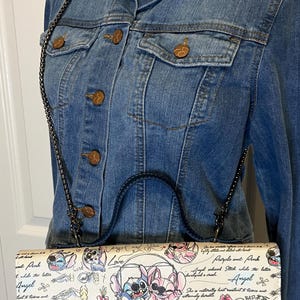 May include: A denim jacket with a black chain strap purse featuring a cartoon design of Stitch and Angel. The purse has a navy blue base and a white top with a colorful illustration and text. The jacket has copper-colored buttons.
