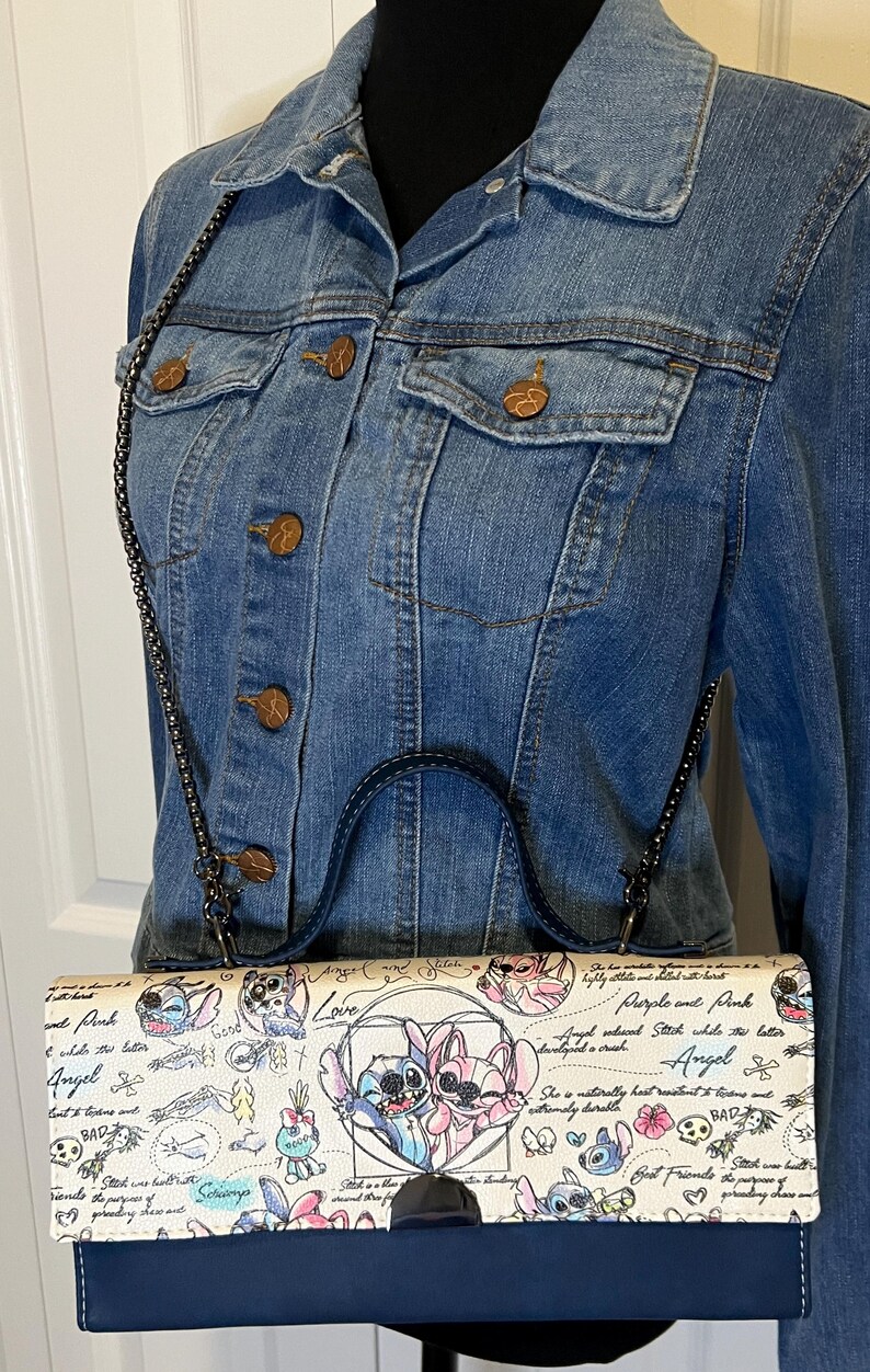 May include: A denim jacket with a black chain strap purse featuring a cartoon design of Stitch and Angel. The purse has a navy blue base and a white top with a colorful illustration and text. The jacket has copper-colored buttons.